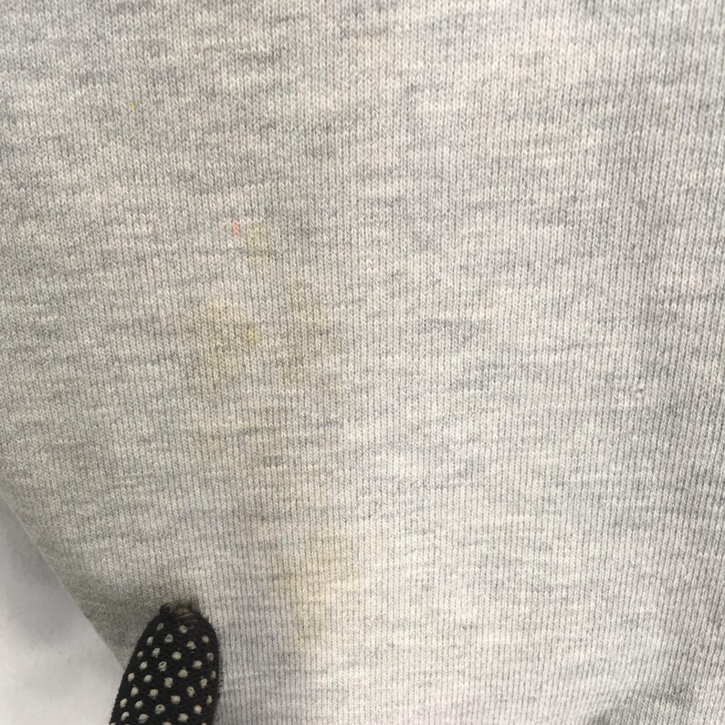May include: Close-up of a light gray fabric with a ribbed texture. The fabric has a few small stains and a dark object with white polka dots is visible at the bottom. The fabric appears to be a knit material.