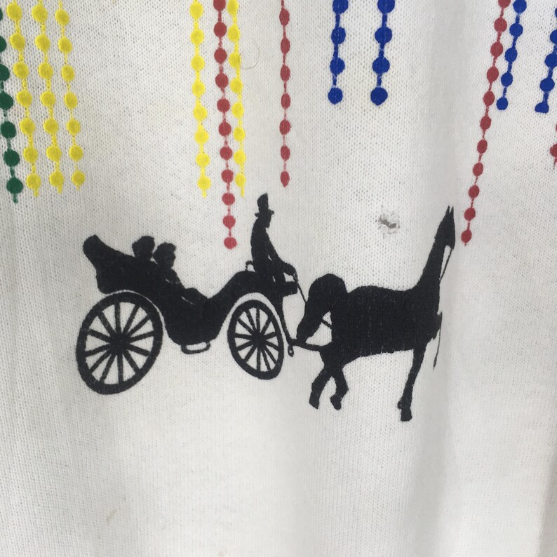 May include: White fabric with a black silhouette of a horse-drawn carriage and passengers. Colorful beaded strands in yellow, red, blue, and green are at the top. The design is likely for a shirt or textile.