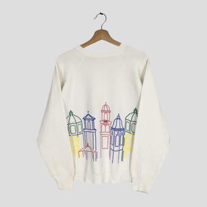 May include: White sweatshirt with a colorful embroidered cityscape design. The design features buildings with domes and towers in shades of blue, red, green, and yellow. The sweatshirt is hanging on a wooden hanger.
