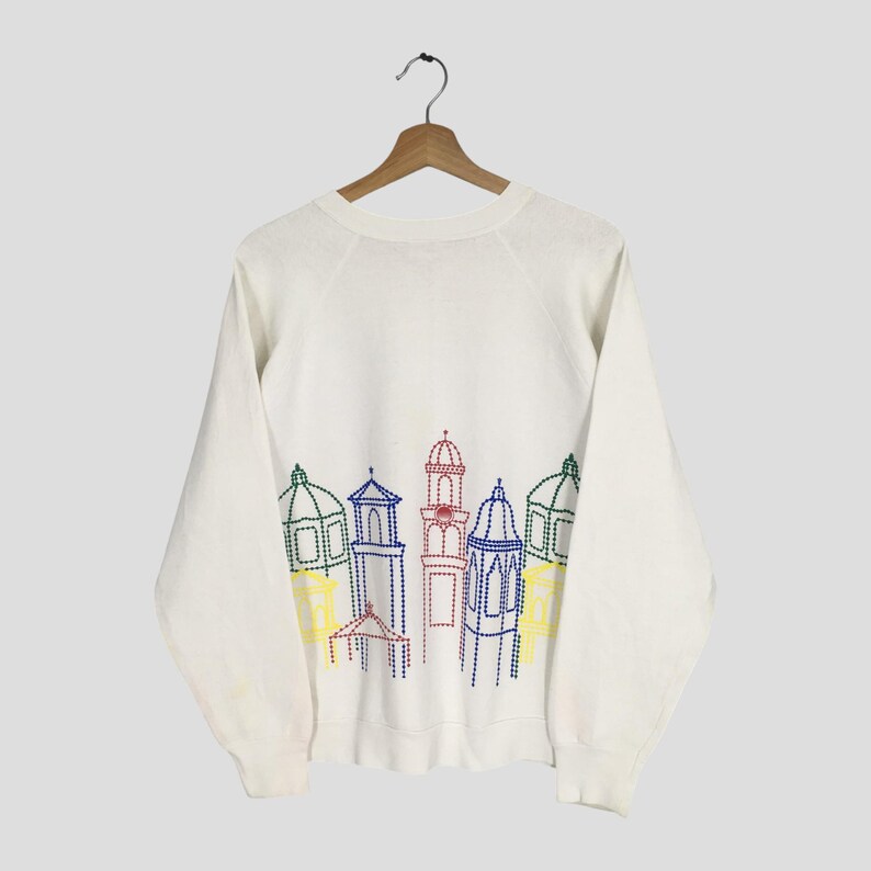 May include: White sweatshirt with a colorful embroidered cityscape design. The design features buildings with domes and towers in shades of blue, red, green, and yellow. The sweatshirt is hanging on a wooden hanger.