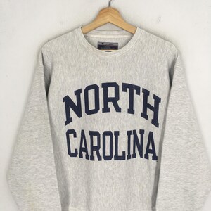 May include: A light gray Champion sweatshirt with the words "NORTH CAROLINA" in navy blue block letters across the chest. The sweatshirt has a crew neck and long sleeves, and is hanging on a wooden hanger.