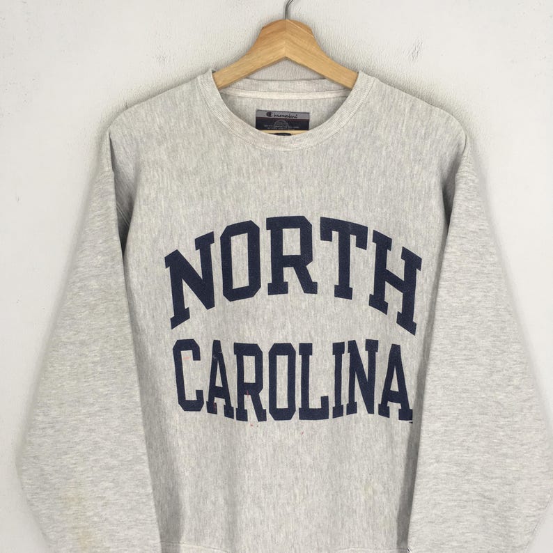 May include: A light gray Champion sweatshirt with the words "NORTH CAROLINA" in navy blue block letters across the chest. The sweatshirt has a crew neck and long sleeves, and is hanging on a wooden hanger.