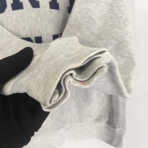 May include: Close-up of a gray sweatshirt sleeve with a ribbed cuff. The fabric shows slight discoloration and a small red stain. Dark blue lettering is visible on the upper portion of the sweatshirt.