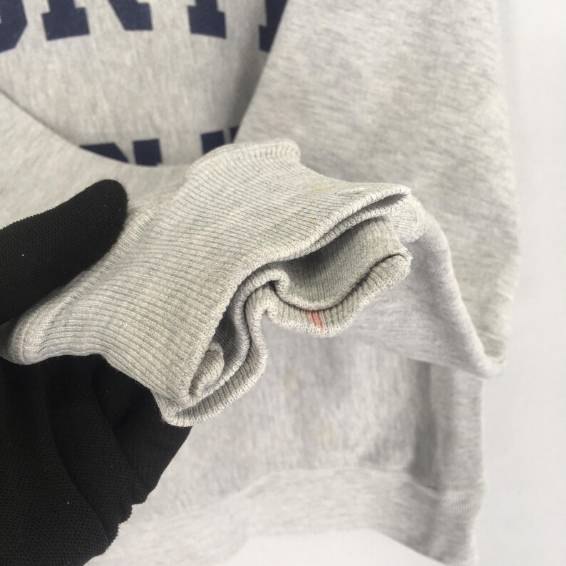 May include: Close-up of a gray sweatshirt sleeve with a ribbed cuff. The fabric shows slight discoloration and a small red stain. Dark blue lettering is visible on the upper portion of the sweatshirt.