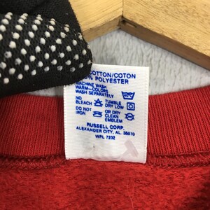 May include: Close-up of a red garment tag with care instructions. The tag reads "COTTON/COTON % POLYESTER" and includes washing symbols. The tag also includes the text "RUSSELL CORP. ALEXANDER CITY, AL 35010 WPL 7232".