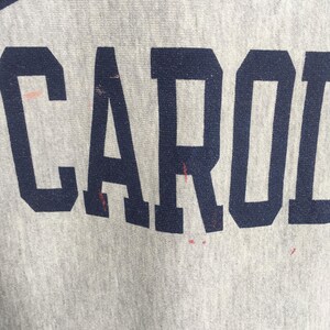 May include: Close-up of a light gray fabric with the word "CAROL" printed in navy blue block letters. The fabric has some red stains. The image is well-lit and the text is clear.