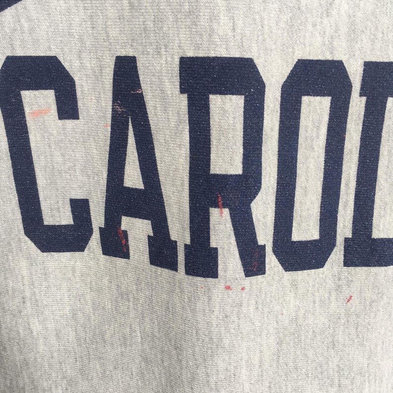 May include: Close-up of a light gray fabric with the word "CAROL" printed in navy blue block letters. The fabric has some red stains. The image is well-lit and the text is clear.