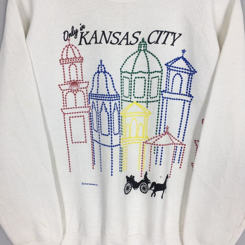 May include: White sweatshirt with a colorful graphic of Kansas City landmarks. The design includes the text "Only in KANSAS CITY" above the buildings, and a horse-drawn carriage at the bottom. The buildings are outlined in various colors.
