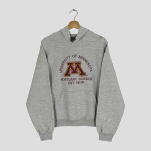 Könnte beinhalten: Ein graumelierter Kapuzenpullover mit einem kastanienbraunen und goldenen Aufdruck University of Minnesota Mortuary Science Est. 1908 auf der Brust. Der Pullover verfügt über eine Kängurutasche vorne, lange Ärmel mit Rippbündchen und eine Kapuze.