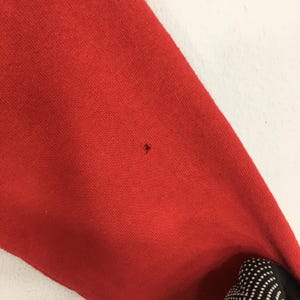 May include: Close-up of a red fabric with a small hole. The fabric appears to be a portion of a garment, possibly a sweatshirt or sweater. The texture of the material is visible, and the color is a vibrant red.