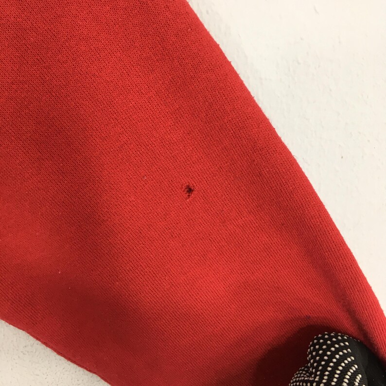 May include: Close-up of a red fabric with a small hole. The fabric appears to be a portion of a garment, possibly a sweatshirt or sweater. The texture of the material is visible, and the color is a vibrant red.