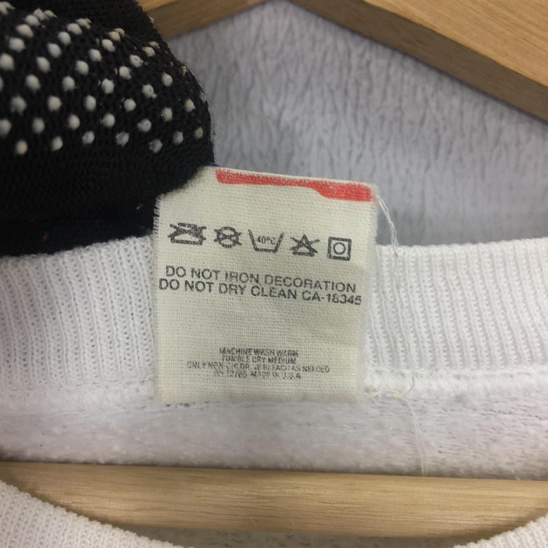 May include: Close-up of a white fabric tag with care instructions. The tag includes symbols for washing, drying, and ironing, along with text indicating the garment should not be ironed or dry cleaned. The tag also has the text "CA-18345".