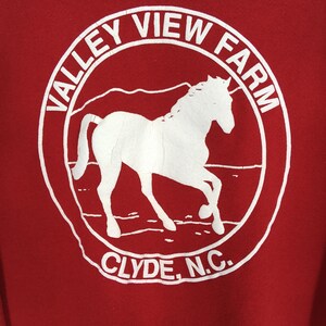 May include: Red sweatshirt with a white circular graphic. The graphic features a horse silhouette inside a circle with the words "VALLEY VIEW FARM" and "CLYDE, N.C." in white text. The horse is in motion.