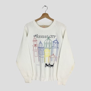 May include: White sweatshirt featuring a colorful embroidered design of Kansas City landmarks. The design includes various buildings in red, blue, yellow, and green, with the text "Kansas City" above the buildings. A horse-drawn carriage is also depicted.