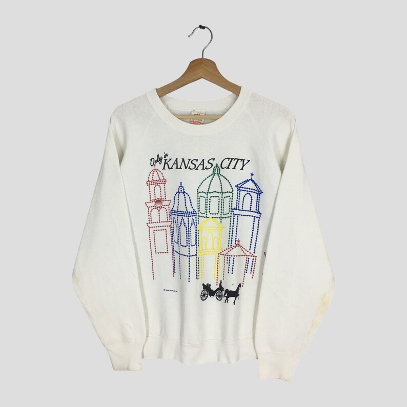 May include: White sweatshirt featuring a colorful embroidered design of Kansas City landmarks. The design includes various buildings in red, blue, yellow, and green, with the text "Kansas City" above the buildings. A horse-drawn carriage is also depicted.