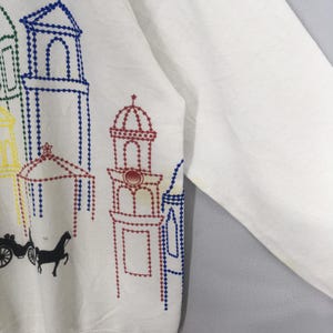 May include: White sweatshirt featuring a colorful embroidered design of buildings, a horse-drawn carriage, and a horse. The embroidery uses blue, green, yellow, and red thread. The sleeve is visible on the right side of the image.