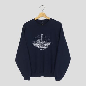 May include: Navy blue sweatshirt featuring a white graphic of a coastal scene with sailboats, buildings, and the text "Cape Cod". The crew neck sweatshirt is made of a soft material and has long sleeves.