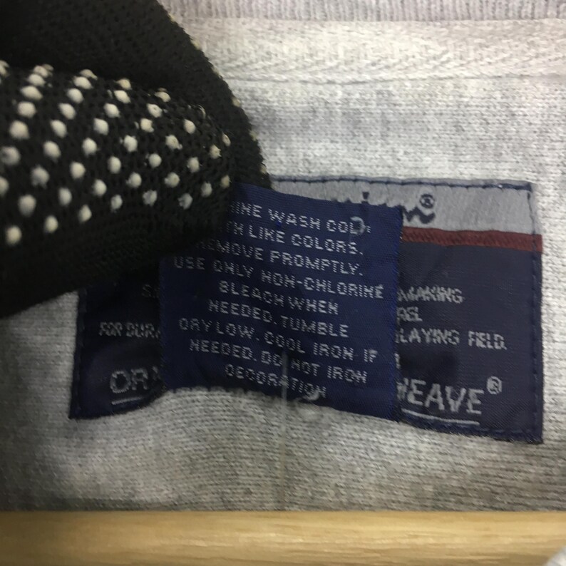 May include: Close-up of a gray fabric with a dark blue tag. The tag has white text with washing instructions and the word "WEAVE". The fabric is slightly textured.