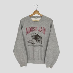 May include: A grey sweatshirt featuring the text "MOOSE JAW" in burgundy above a moose graphic. Below the moose graphic is the text "SASKATCHWAN, CANADA". The sweatshirt is suspended from a wooden hanger.
