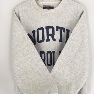 May include: A light gray Champion sweatshirt with the words "NORTH CAROLINA" in navy blue block letters. The crew neck sweatshirt has long sleeves and a tag at the back of the neck.