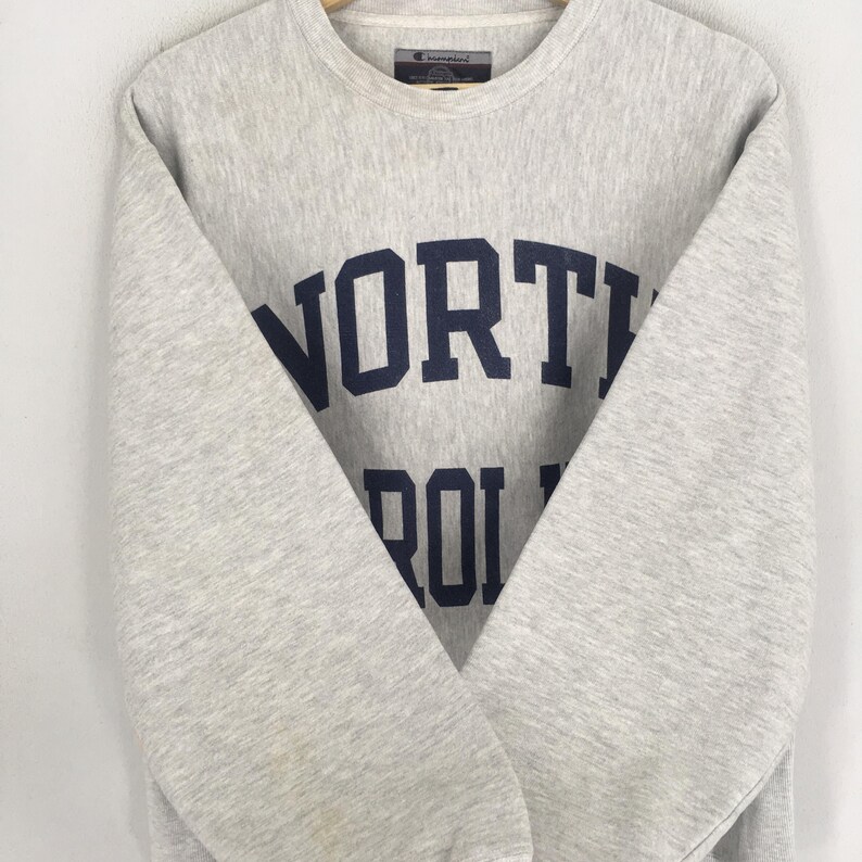 May include: A light gray Champion sweatshirt with the words "NORTH CAROLINA" in navy blue block letters. The crew neck sweatshirt has long sleeves and a tag at the back of the neck.