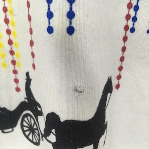 May include: White fabric with a black illustration of a horse-drawn carriage and a horse. Colorful beaded strings in red, yellow, and blue are also present. The fabric has a small hole.