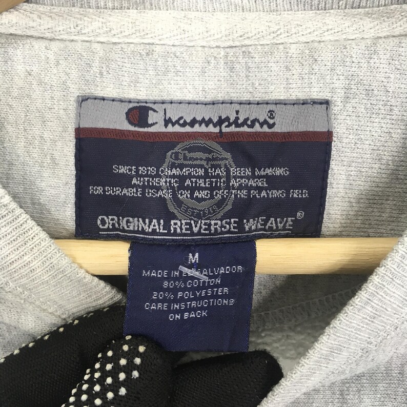 May include: Close-up of a gray Champion sweatshirt tag. The tag is dark blue with white text that reads "ORIGINAL REVERSE WEAVE" and "AUTHENTIC ATHLETIC APPAREL". The tag also includes the Champion logo and the size "M".