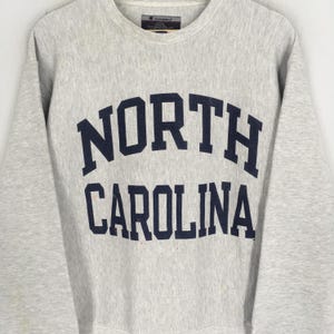 May include: A light gray Champion sweatshirt with the words "NORTH CAROLINA" in navy blue block letters across the chest. The sweatshirt has a crew neck and long sleeves.