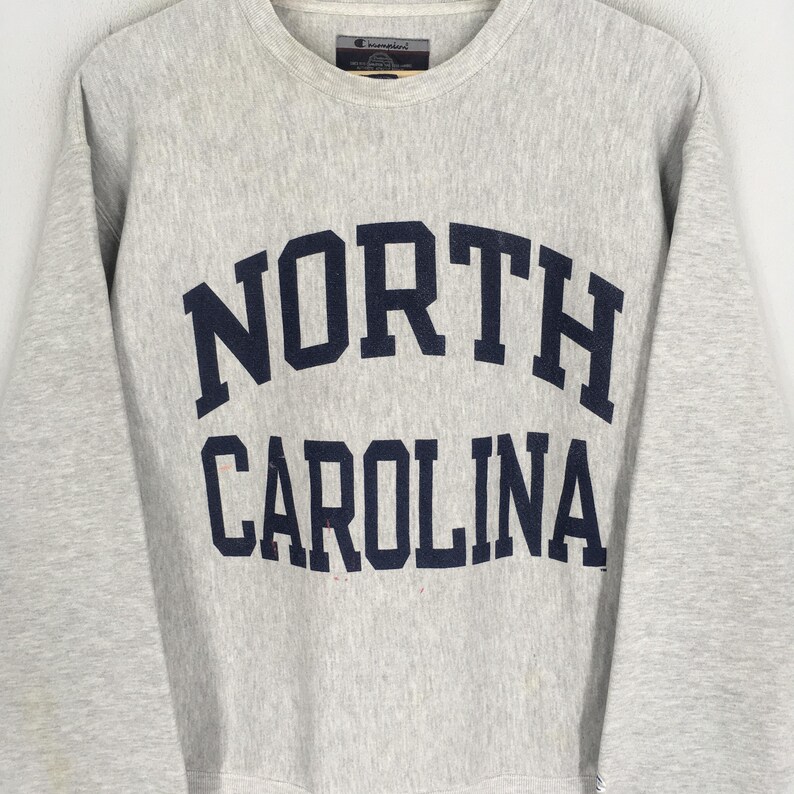 May include: A light gray Champion sweatshirt with the words "NORTH CAROLINA" in navy blue block letters across the chest. The sweatshirt has a crew neck and long sleeves.