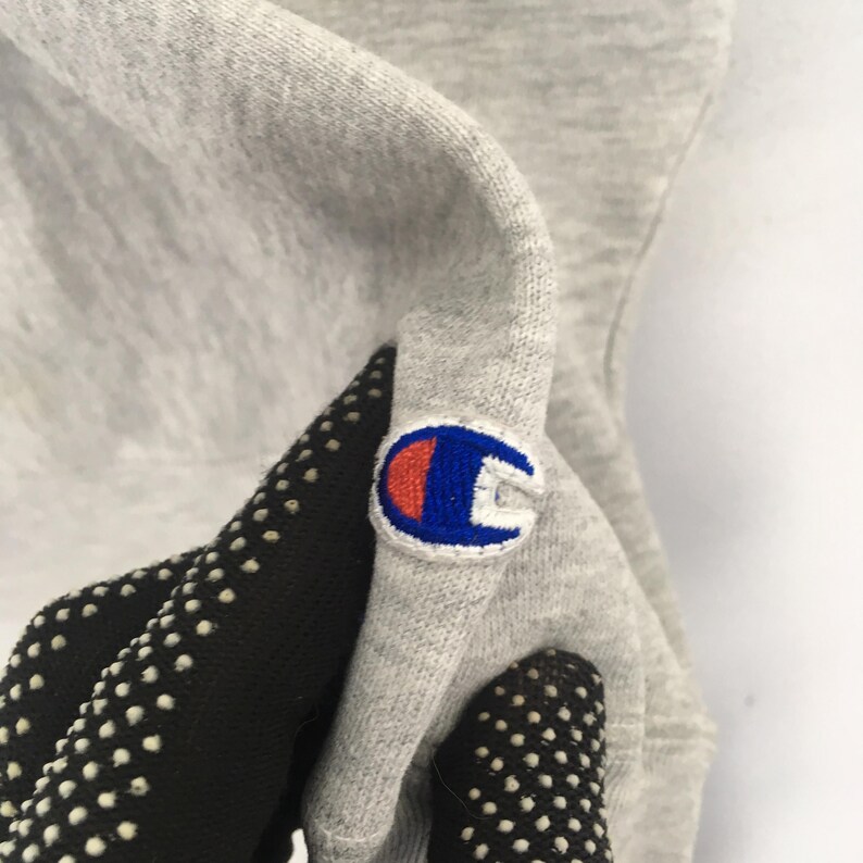 May include: Close-up of a light gray Champion sweatshirt. The Champion logo, a white, blue, and red patch, is sewn on the sleeve. The sleeve is held by a black glove with white dots.