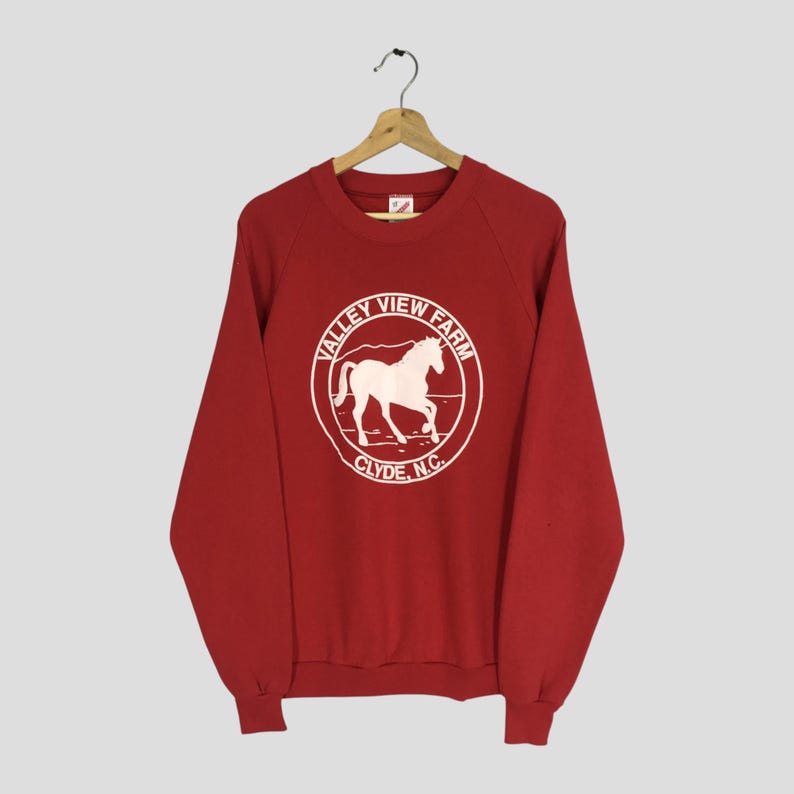 May include: Red crewneck sweatshirt with a white graphic of a horse inside a circle that reads "Valley View Farm Clyde, N.C.". The sweatshirt is hanging on a wooden hanger.