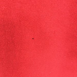 May include: Close-up of a solid red knit fabric. The fabric has a textured appearance with visible knit lines. A small dark spot is visible on the fabric. The image highlights the texture and color of the material.