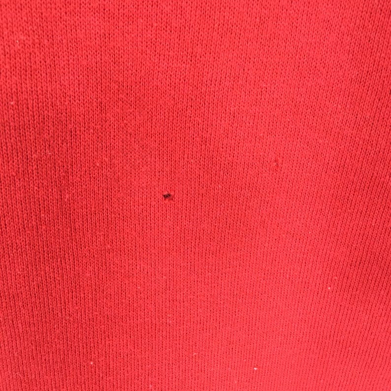 May include: Close-up of a solid red knit fabric. The fabric has a textured appearance with visible knit lines. A small dark spot is visible on the fabric. The image highlights the texture and color of the material.