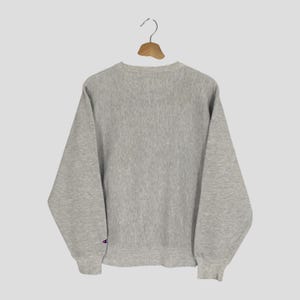 May include: A heather gray Champion sweatshirt hangs from a wooden hanger. The crew neck sweatshirt has long sleeves and a small Champion logo on the left sleeve cuff. The sweatshirt is made of a soft, textured fabric.
