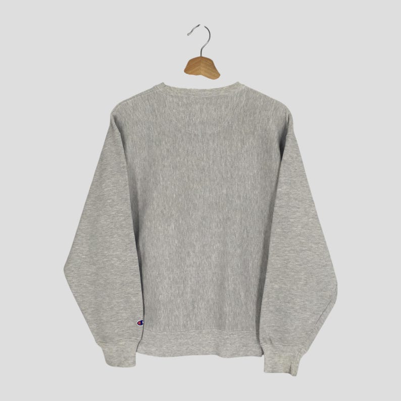 May include: A heather gray Champion sweatshirt hangs from a wooden hanger. The crew neck sweatshirt has long sleeves and a small Champion logo on the left sleeve cuff. The sweatshirt is made of a soft, textured fabric.