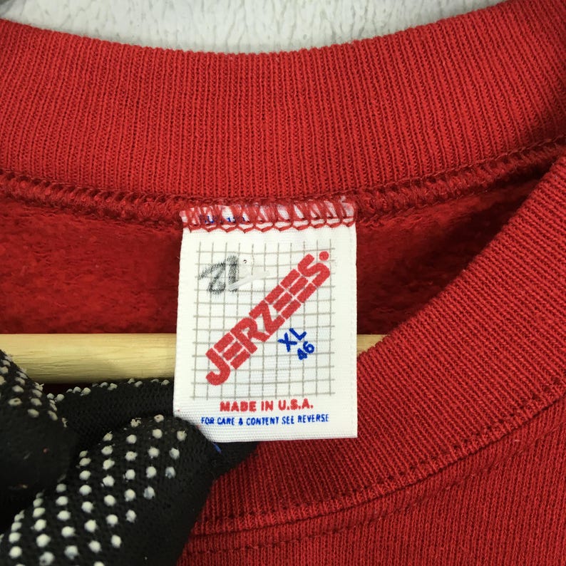 May include: Close-up of a red sweatshirt collar with a white tag. The tag reads "JERZEES" in red letters, with "XL 46" in blue. The tag also states "MADE IN U.S.A." and "FOR CARE & CONTENT SEE REVERSE".
