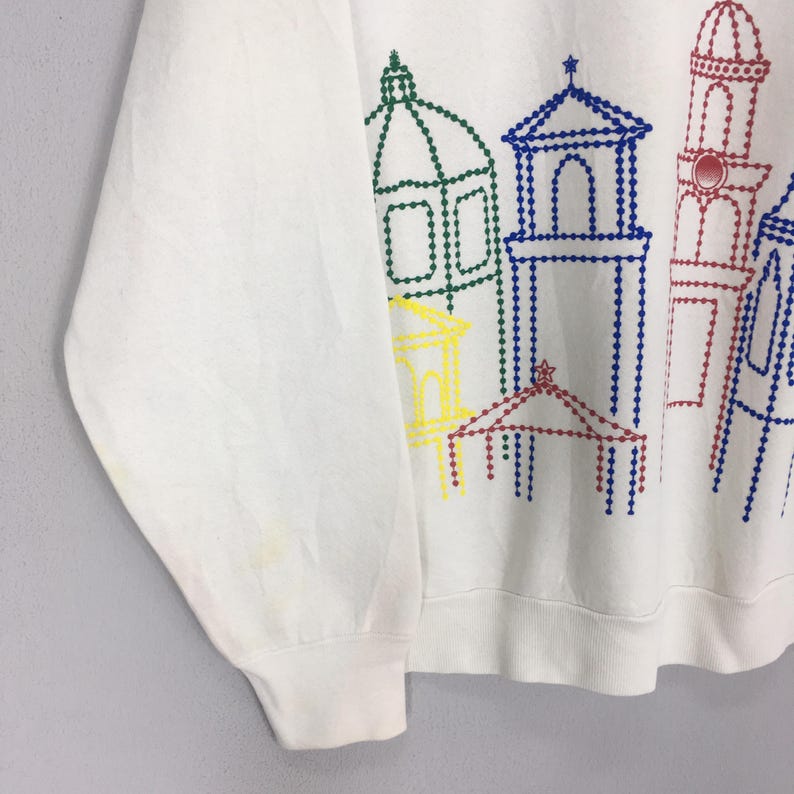 May include: White sweatshirt featuring a colorful, dotted line illustration of buildings. The design includes green, blue, red, and yellow architectural elements. The sweatshirt has a crew neck and long sleeves.