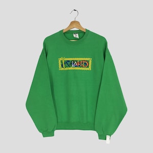 Puede incluir: Sudadera verde con la palabra "Ireland" bordada en un diseño rectangular. El bordado presenta colores dorados, azules, rojos y verdes. La sudadera tiene mangas largas y cuello redondo.