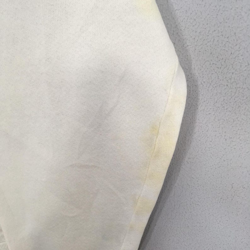 May include: Close-up of a white fabric with visible stains. The fabric appears to be a knit or woven material, with a slightly textured surface. The stains are yellow and concentrated along the edge of the fabric.