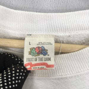 May include: Close-up of a white Fruit of the Loom sweatshirt tag. The tag is rectangular with the brand name and a graphic of fruit. The tag also indicates the size is L and the material is 50% cotton and 50% polyester. Made in the U.S.A.