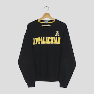 Pode incluir: Camisola preta de gola redonda com a palavra "APPALACHIAN" em letras amarelas no peito. Um logótipo "A" amarelo está no canto superior direito. A camisola está pendurada num cabide de madeira.