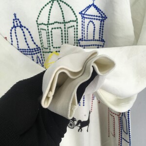 May include: White sweatshirt with embroidered architectural designs in blue, green, and red. The cuff is white with a black and white striped interior. A black cat pulling a carriage is also embroidered on the sweatshirt.