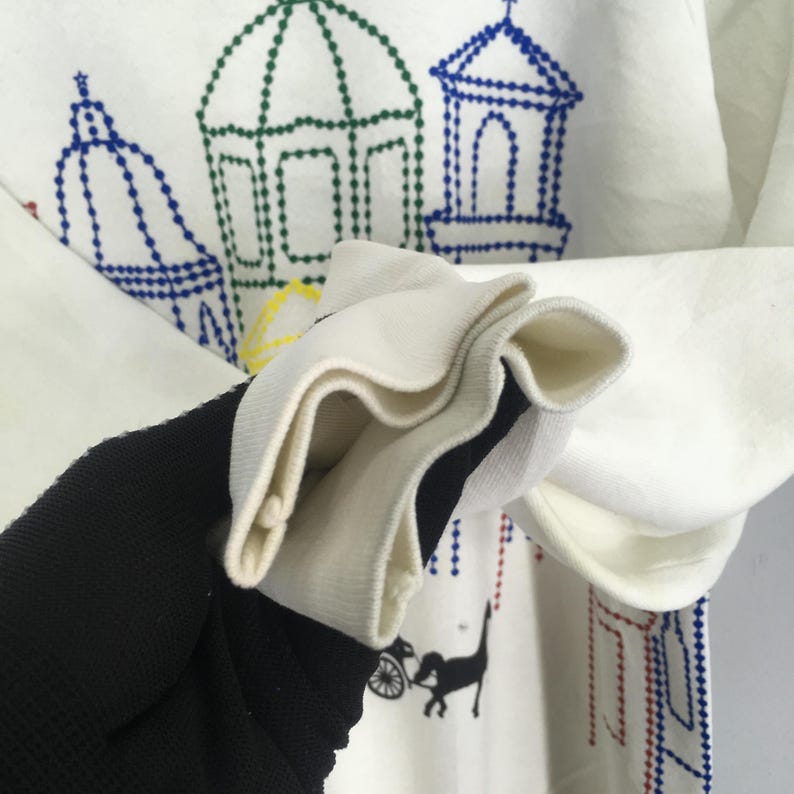 May include: White sweatshirt with embroidered architectural designs in blue, green, and red. The cuff is white with a black and white striped interior. A black cat pulling a carriage is also embroidered on the sweatshirt.
