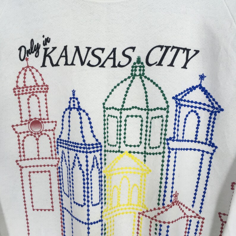 May include: White sweatshirt featuring the text "Only in KANSAS CITY" above a colorful dotted outline of city buildings. The buildings are outlined in red, blue, green, and yellow.