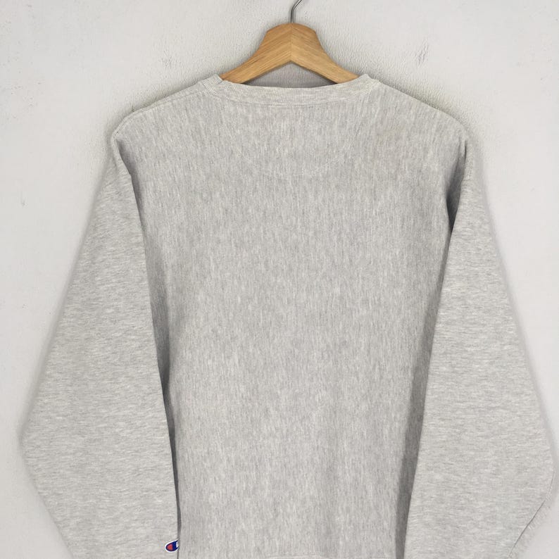 May include: A light gray Champion sweatshirt with a crew neck, hanging on a wooden hanger. The Champion logo is visible on the left sleeve. The sweatshirt is made of a soft, comfortable material.