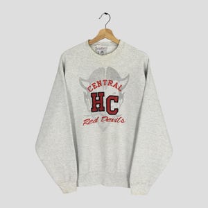 May include: A light grey sweatshirt featuring the text "CENTRAL HC Red Devils" in red and grey. The sweatshirt is displayed on a wooden hanger. The crew neck sweatshirt is against a light grey backdrop.