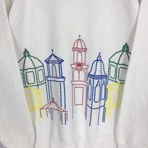 May include: White sweatshirt featuring a colorful embroidered design of stylized buildings. The buildings are outlined in green, blue, red, and yellow thread, creating a vibrant and eye-catching pattern. The sweatshirt is a casual, comfortable garment.