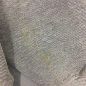 May include: Close-up of a gray sweatshirt fabric with visible stains. The fabric has a ribbed texture and appears to be made of a soft material. The stains are yellowish-brown and scattered across the surface.
