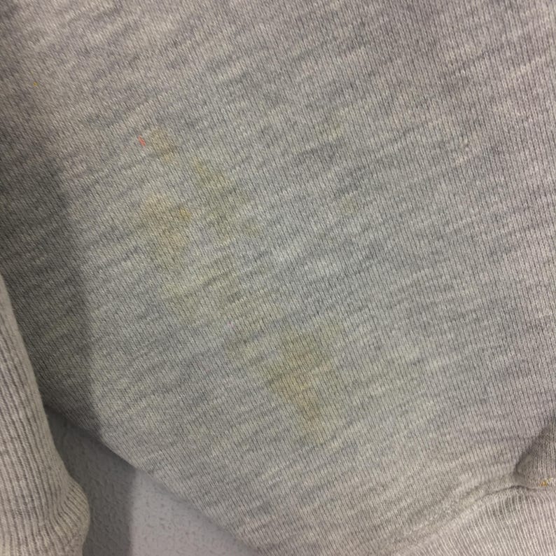 May include: Close-up of a gray sweatshirt fabric with visible stains. The fabric has a ribbed texture and appears to be made of a soft material. The stains are yellowish-brown and scattered across the surface.