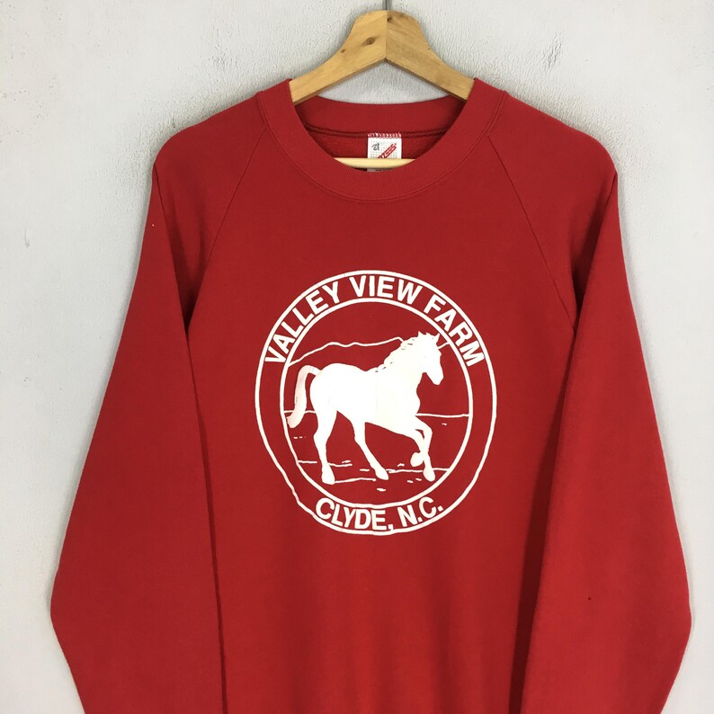 May include: A red sweatshirt with a white circular graphic featuring a horse silhouette and the text "VALLEY VIEW FARM CLYDE, N.C.". The long-sleeved top is hanging on a wooden hanger.